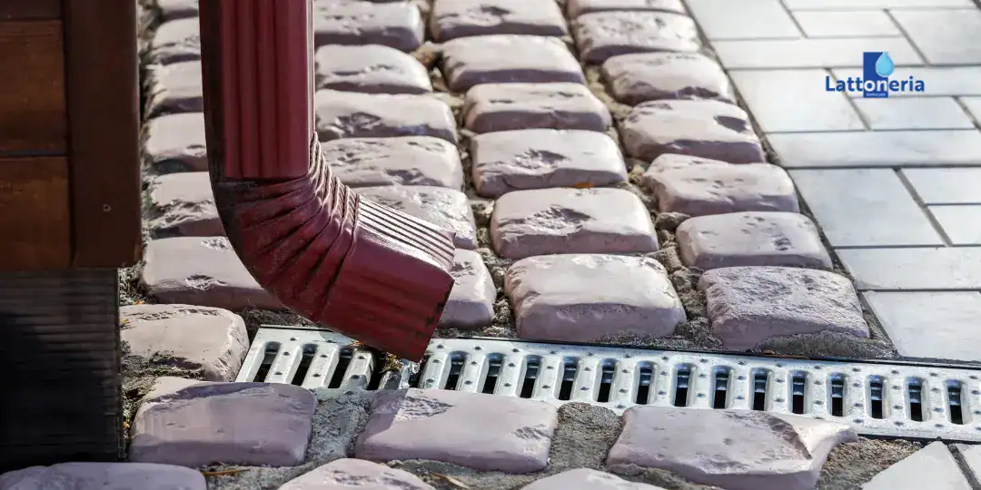 Tubo pluviale in lamiera verniciata che convoglia l’acqua piovana verso una griglia di drenaggio a pavimento, installazione esterna su pavimentazione in pietra.